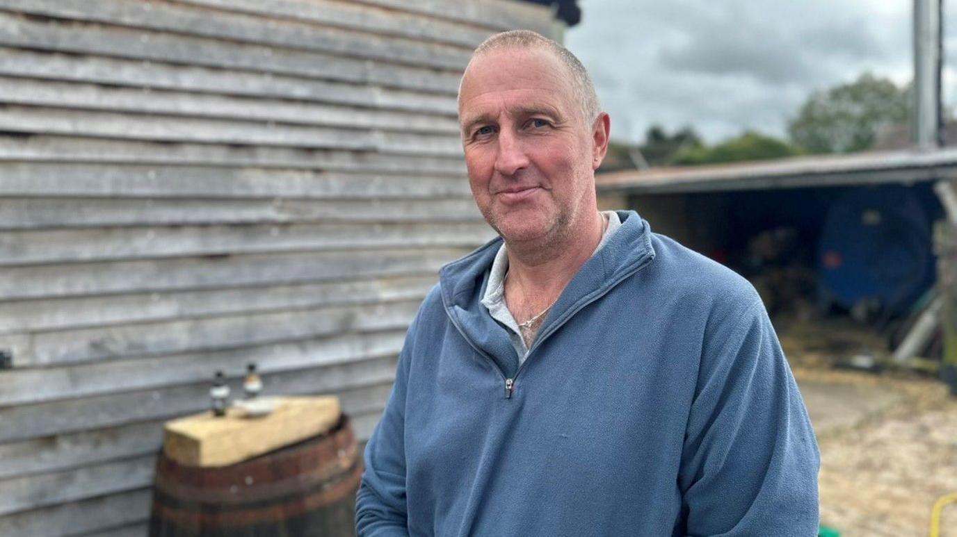 Tom Tame has very short light brown hair. He is wearing a blue zip jumper and is standing in front of a farm building