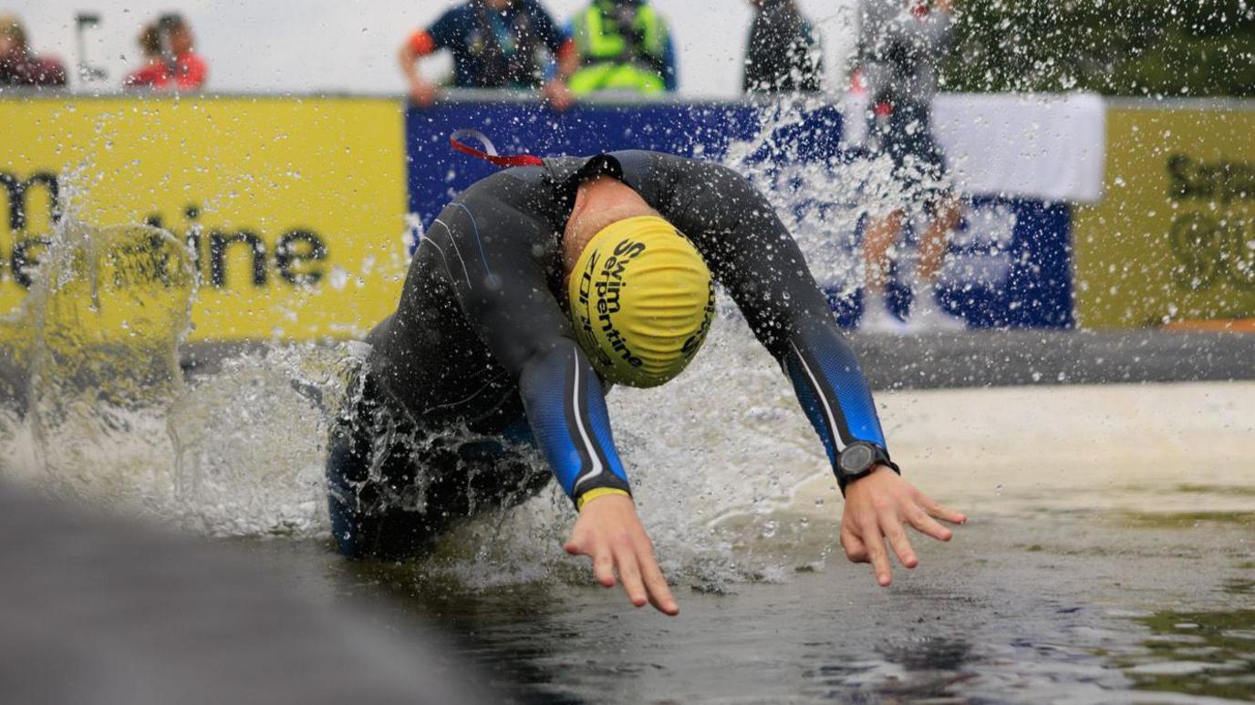 Man in blue wetsuit with  yellow swim cap diving into open water. There are people watching on in the background and trees. There is a yellow and blue Swim Serpentine banner. 