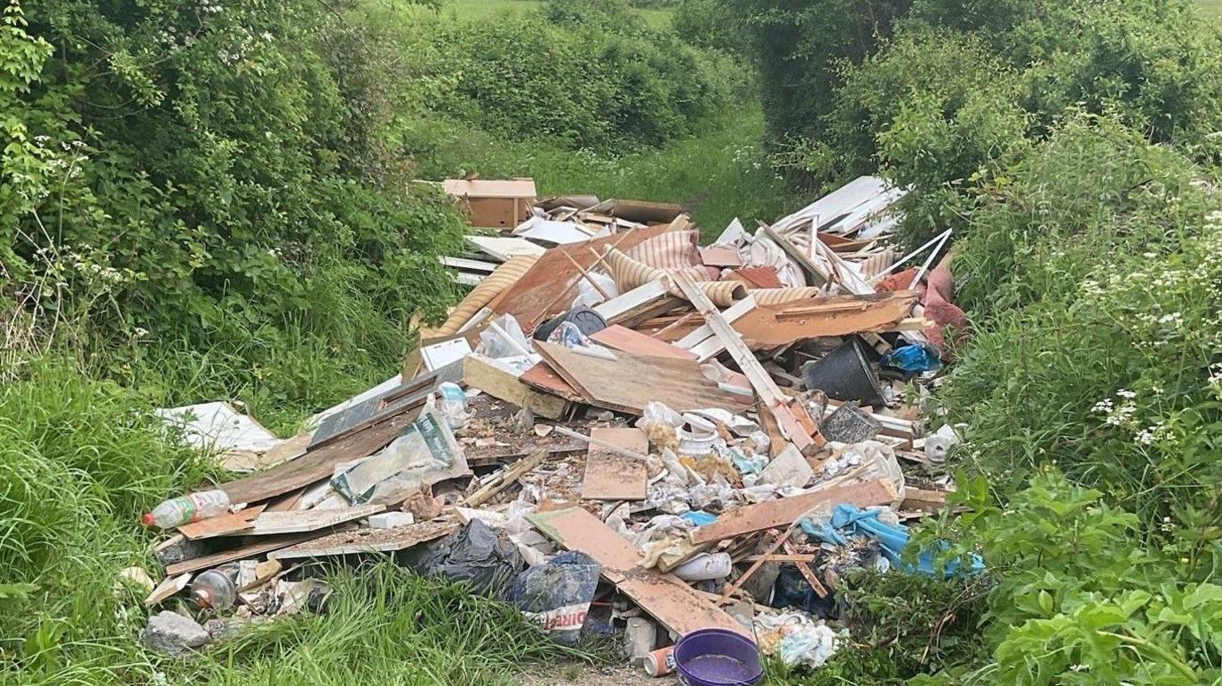 A large amount of construction waste, wood and furnishings in the middle of a grassy path surrounded by green bushes and grass.