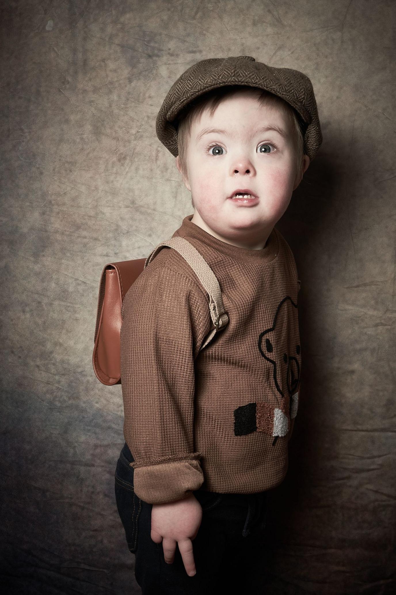 Albie, a young boy, wears a brown flat camp and brown jumper with a teddy bear design on it with a satchel on his back. 