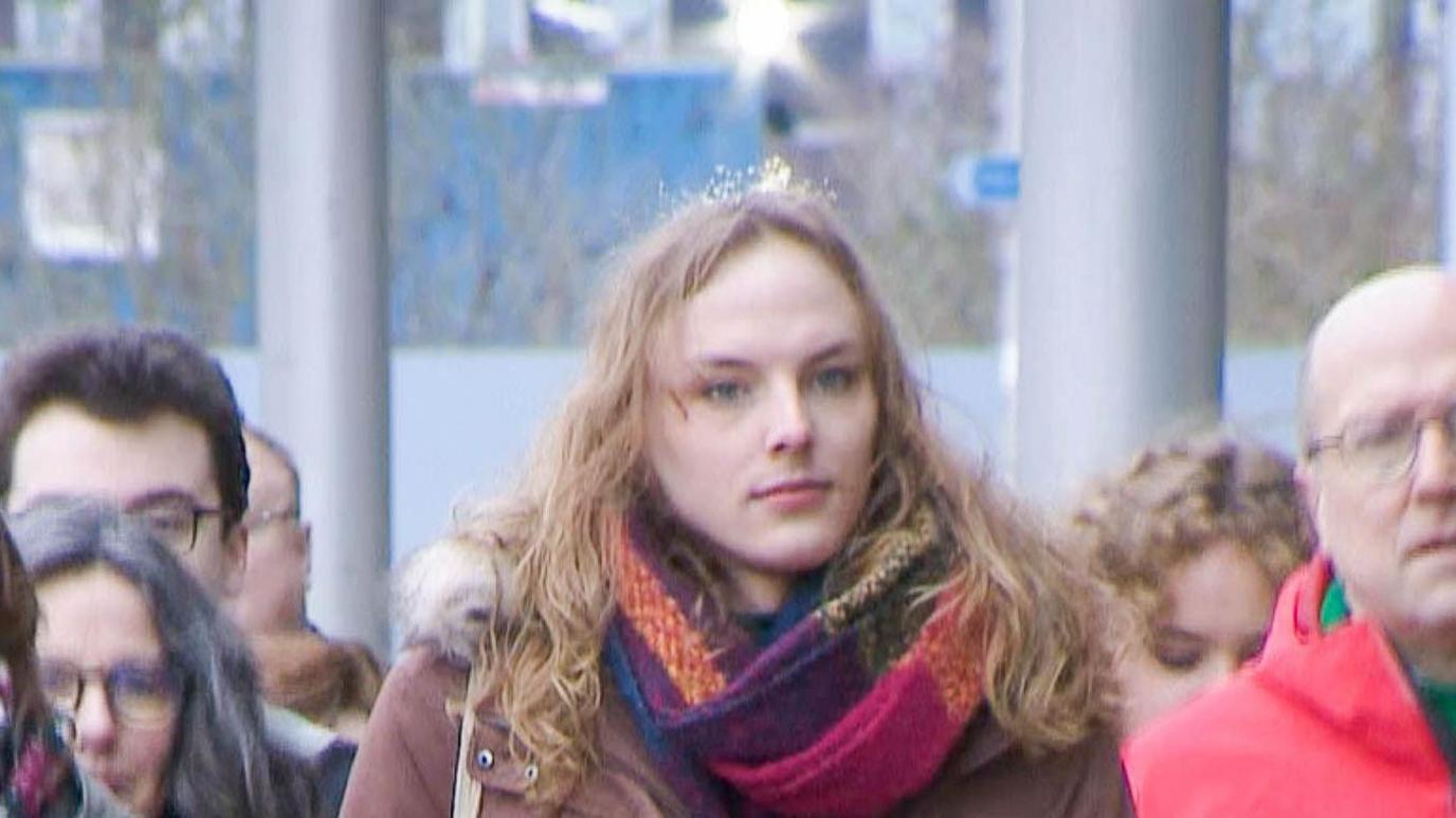 Dr Beth Upton - a trans woman with long hair - walks towards a hearing while wearing a maroon coat and multi coloured scarf.