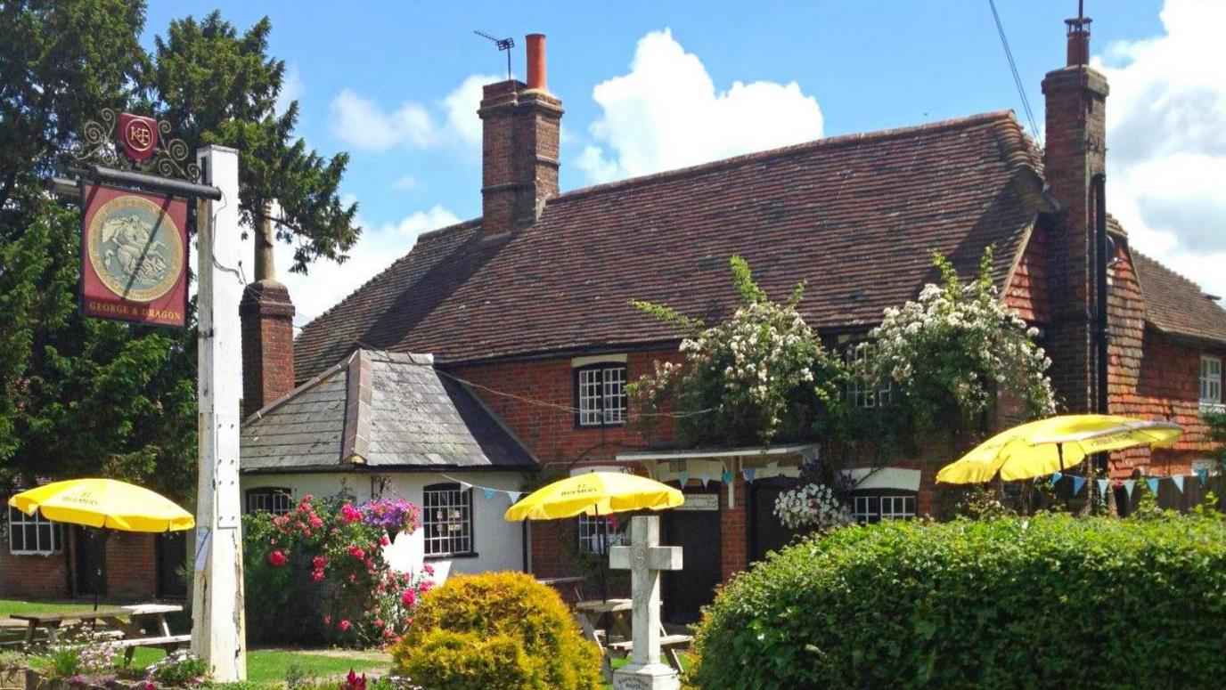 The George and Dragon pub in Dragons Green, West Sussex. The pub is a red bricked building with a white front extension. There is a sign for the pub in the image, alongside three yellow pub umbrellas.