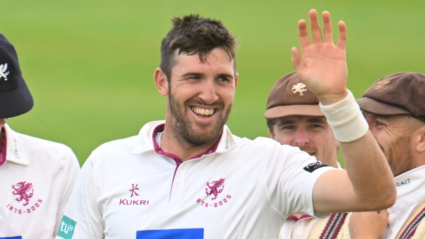 Craig Overton celebrates taking a wicket. He is smiling and holding his hand up.
