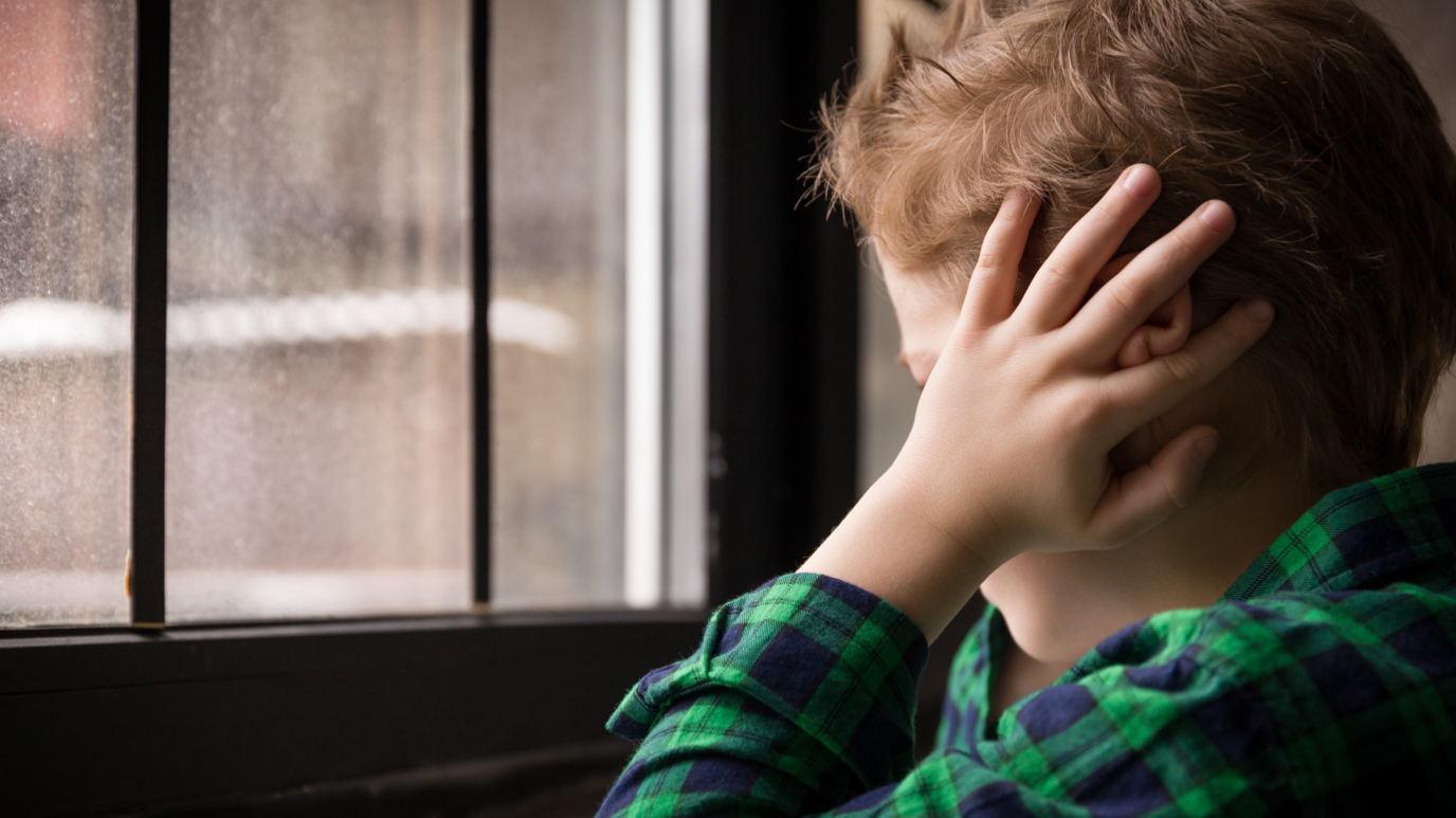 Stock image of a child in a green and blue check shirt staring out of the window with their hands over their ears.  