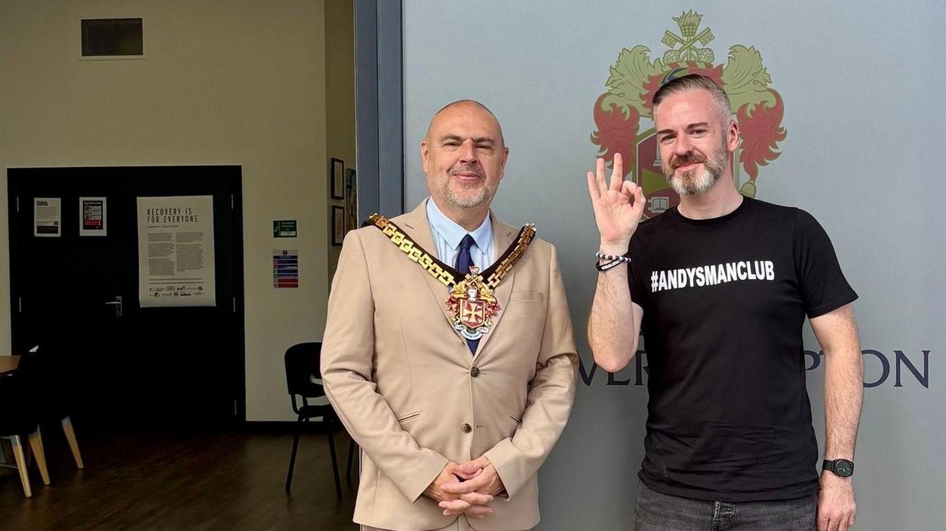 Danny Rice, a man wearing a black t-shirt with the hashtag #andysmanclub in white letter on it, is standing next to the mayor of Wolverhampton Craig Collingswood at The Hub at Wolverhampton railway station. The mayor is wearing a beige suit with the chains of office and a light-coloured shirt and dark blue tie.
