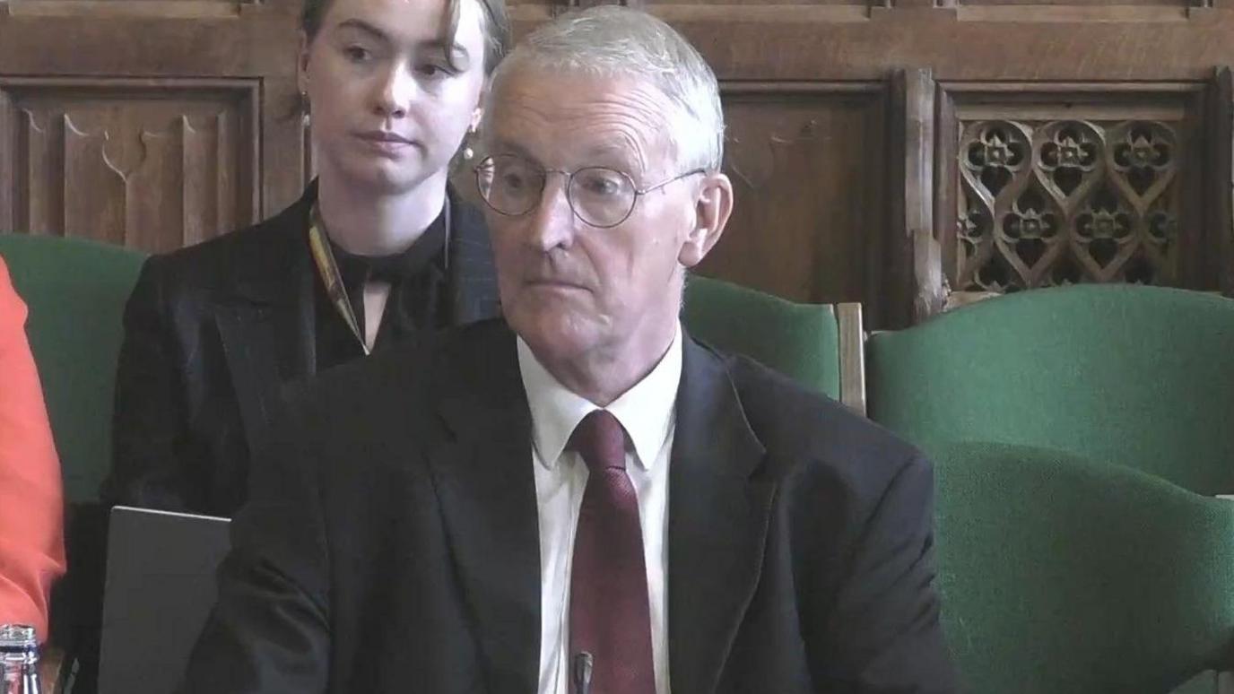 A still of Hilary Benn appearing at the Northern Ireland Affairs Committee in Westminster. He, as well as other attendees, are sitting in green fabric chairs. Benn is an older man with grey/white short hair and a pair of circular metal glasses. He is wearing a black suit jacket, a red tie and a white collared shirt.