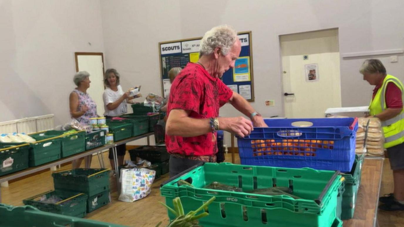 A man in a red t shirt moves produce with his hands from one food crate to another. He is around 70 years old and wearing a red t shirt. Other volunteers can be seen working on tressle tables behind him.