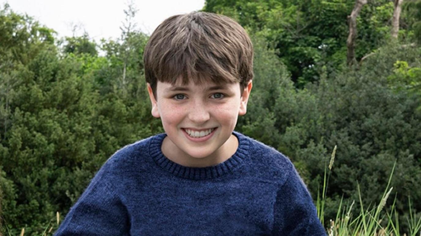 A young boy sitting on the grass in front of green trees. He is wearing a blue jumper and is smiling at the camera.