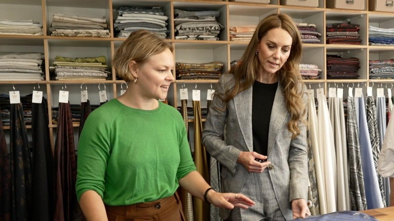 A woman with short blonde hair, wearing a green long sleeved top and brown trousers looks at a table with silk fabric on it. The Princess of Wales listens and looks at the table. She has long brown hair that is lightly curled and she wears a grey suit with a black top underneath.