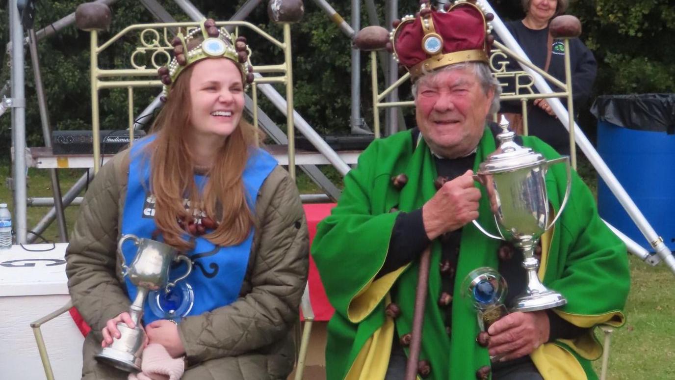 Two people are sat on thrones decorated with conkers. On the left is Kelci Banschbach, she has long light brown hair and is wearing a long dark olive winter coat. She has a crown on her head decorated with conkers. Next to her is an older man in a green and yellow cloak, he also has a conker themed crown and throne. Both smile with their trophies.