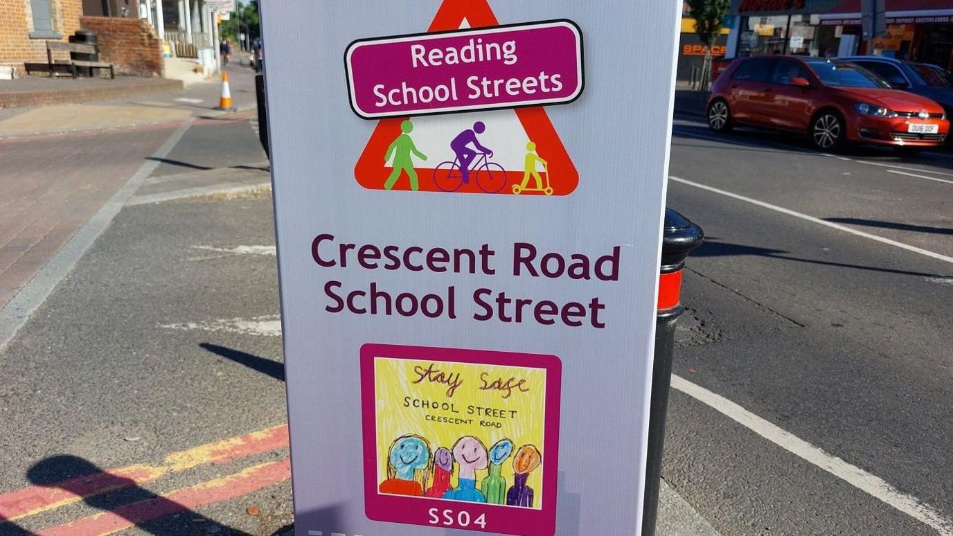 A sign attached to a bollard, which reads "Reading school streets" and "Crescent Road school street". On the sign there is also a child's drawing of several smiling people, which reads "stay safe". Behind the sign, there is a black and red bollard. The sign is on a residential road and there are cars in the background.