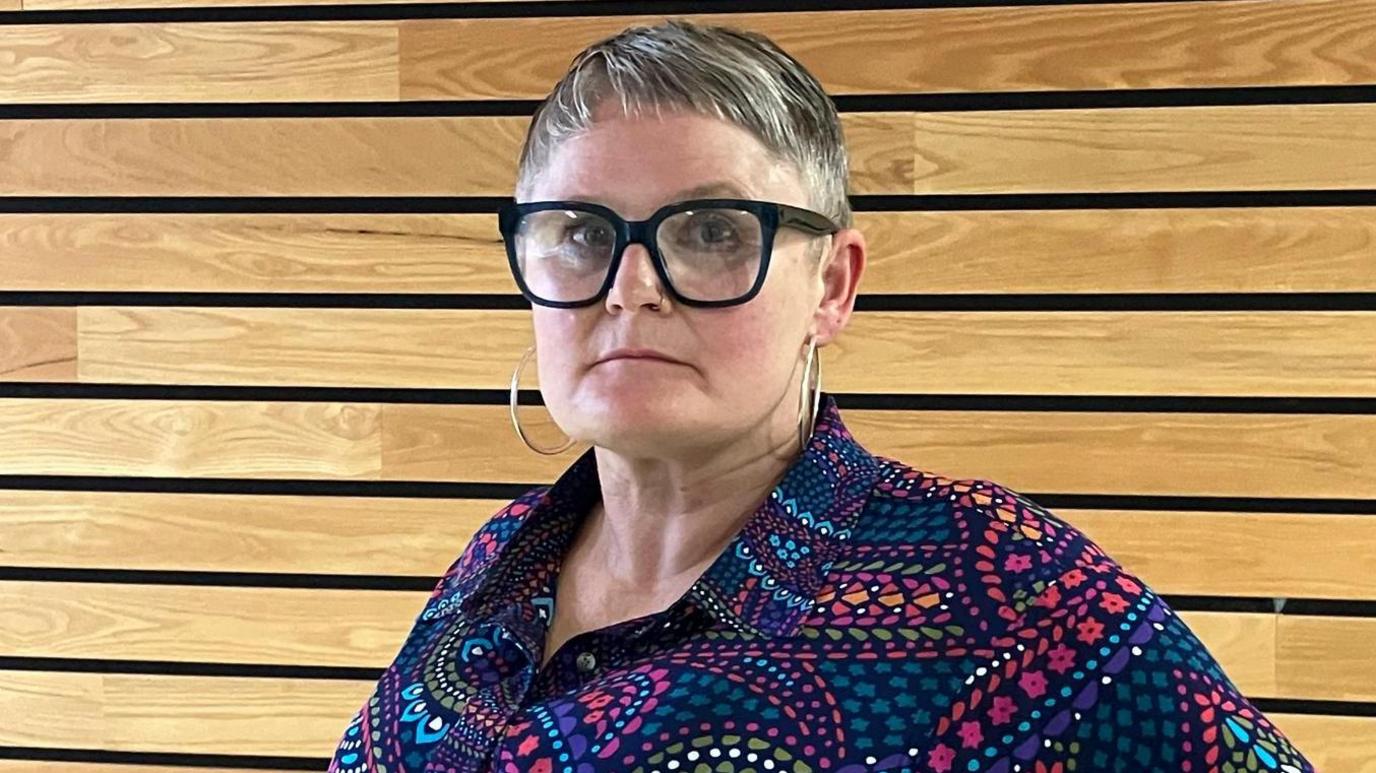 Mae gan Joe William sbectol gyda ffram ddu, gwallt byr ac mae'n gwisgio crys lliwgar. Mae'n edrych mewn i gyfeiriad y camera.