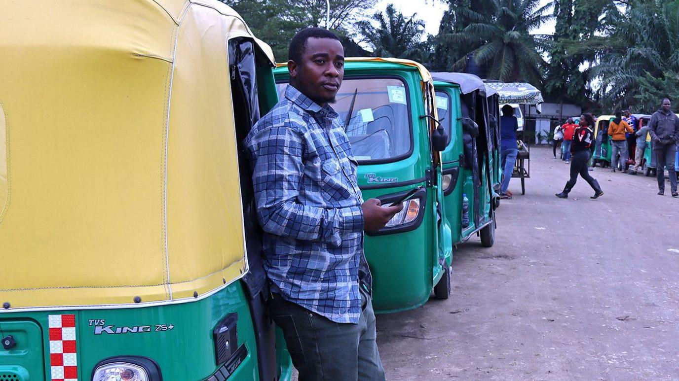 A man in jeans, sliders and a checked shirt stands by his three-wheeled vehicle as he waits to fill up with CNG.