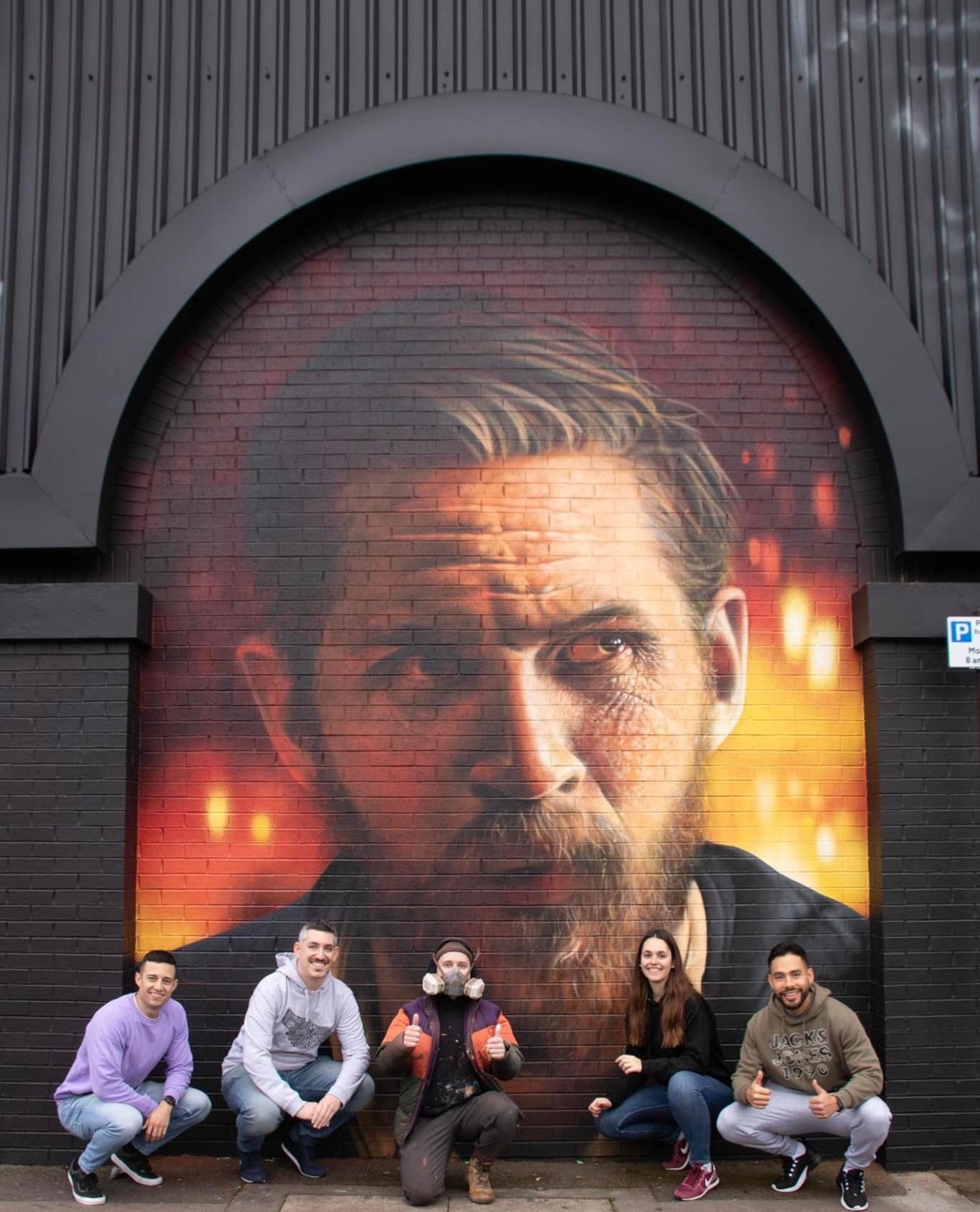 Five people are standing in front of the mural of Alfie Solomons, which has an orange and red background. Tom has brown hair and a beard. 