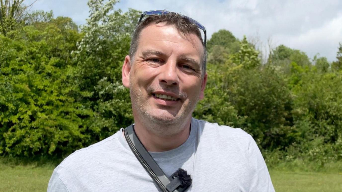 Daniel Baptist in grey t shirt smiling at camera on a sunny day in front of trees and bushes