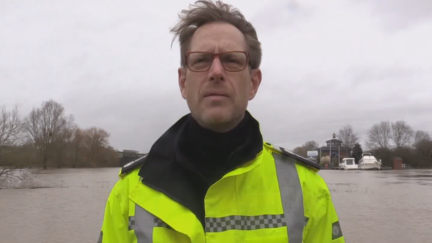Mark Sitton-Kent with brown hair and red-framed glasses, wearing a high vis jacket and stood in front of a raised river.