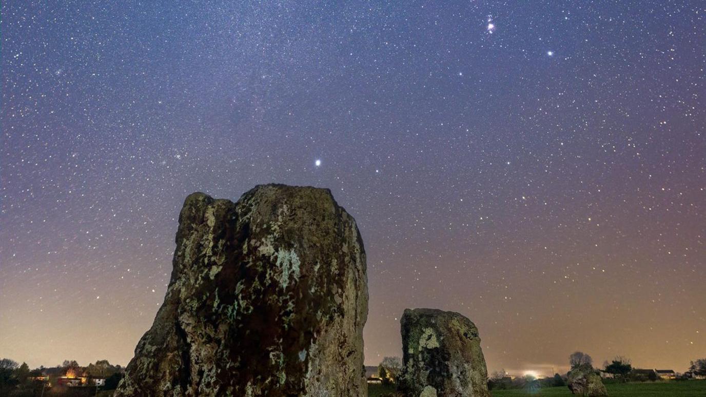 Stanton Drew Stone Circles are photographed at night with stars visible. The sky is blue, purple and orange. The stones are in the foreground.