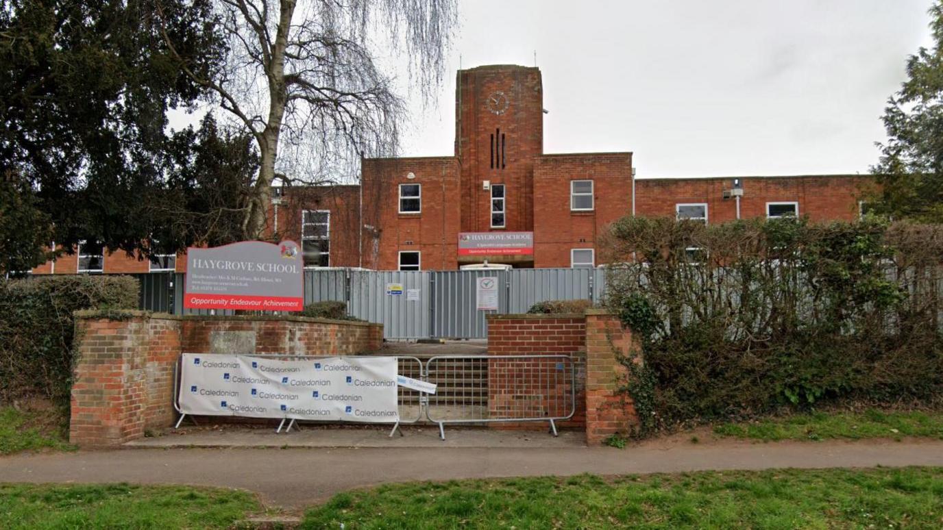 A street view of the entrance to Haygrove School. It is a large brick building with a tall middle section that has a clock on it. The entrance to the school is blocked off by metal fencing and corrugated walls as if under construction.