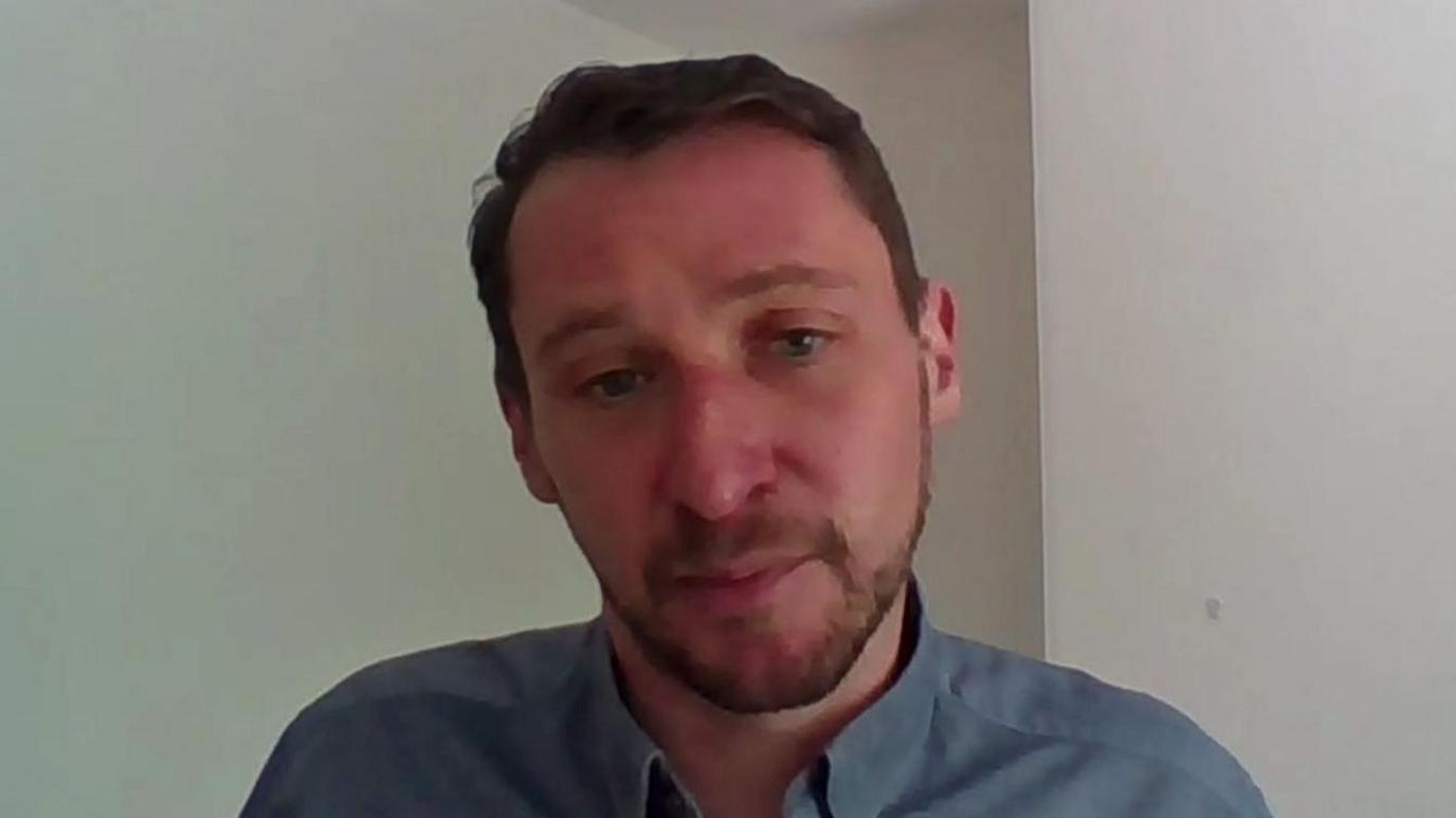 Mr O'Hara is looking at the camera during a Zoom-style interview. He's wearing a grey shirt and the walls behind him are bear and off-white. He has dark hair and a short moustache and beard.