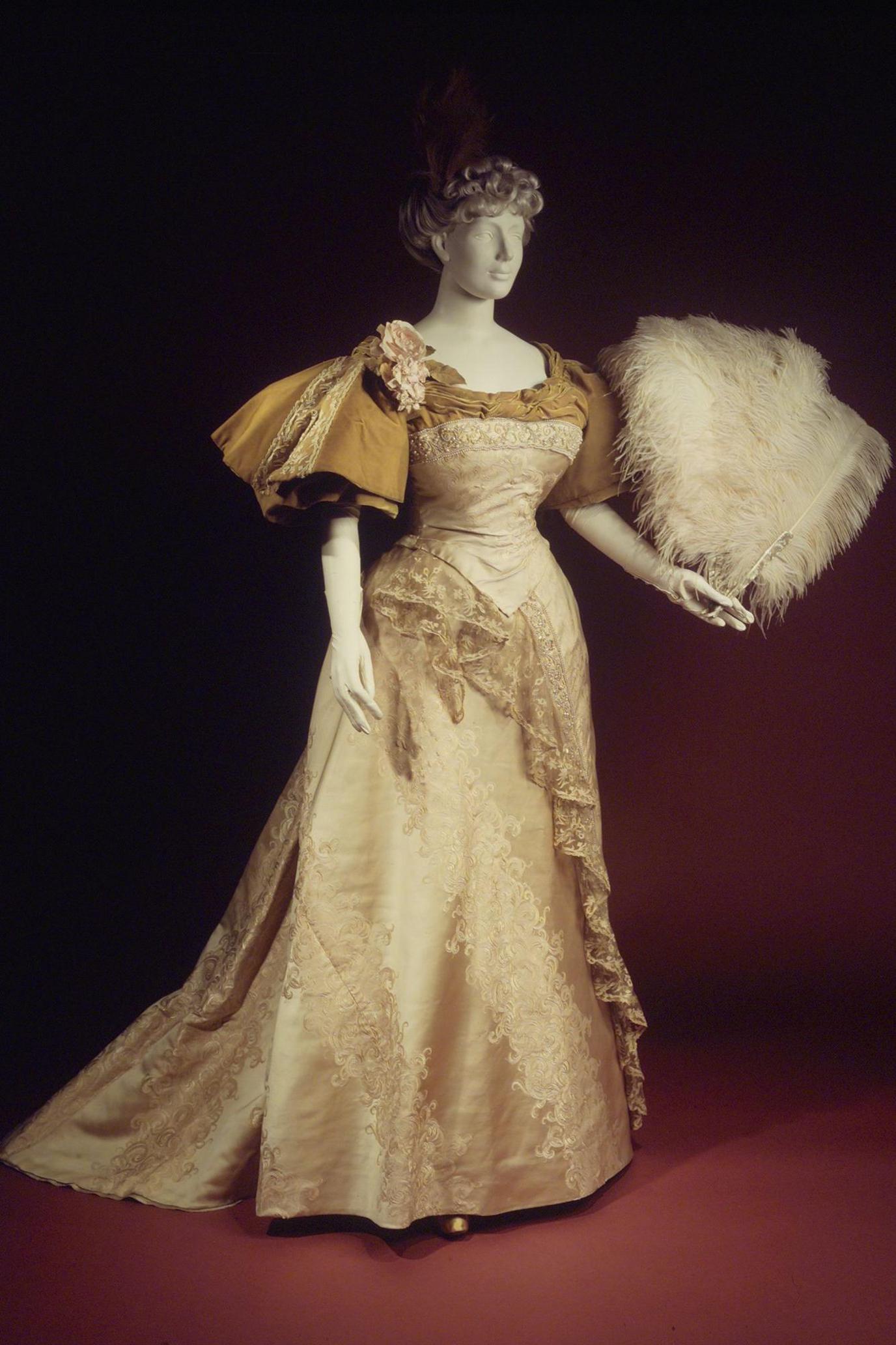 A mannequin is dressed in an evening dress dated from 1894. The dress is gold coloured with a trimming of brown lace and silk. The mannequin is holding a feather fan.