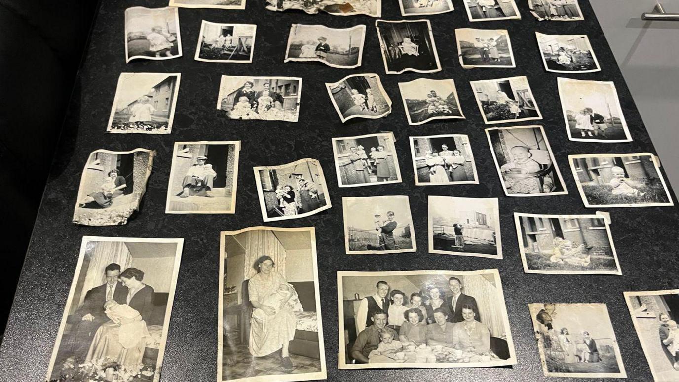 Family photos are spread out across a kitchen table, showing black and white images of children and families as well as a few colour images.