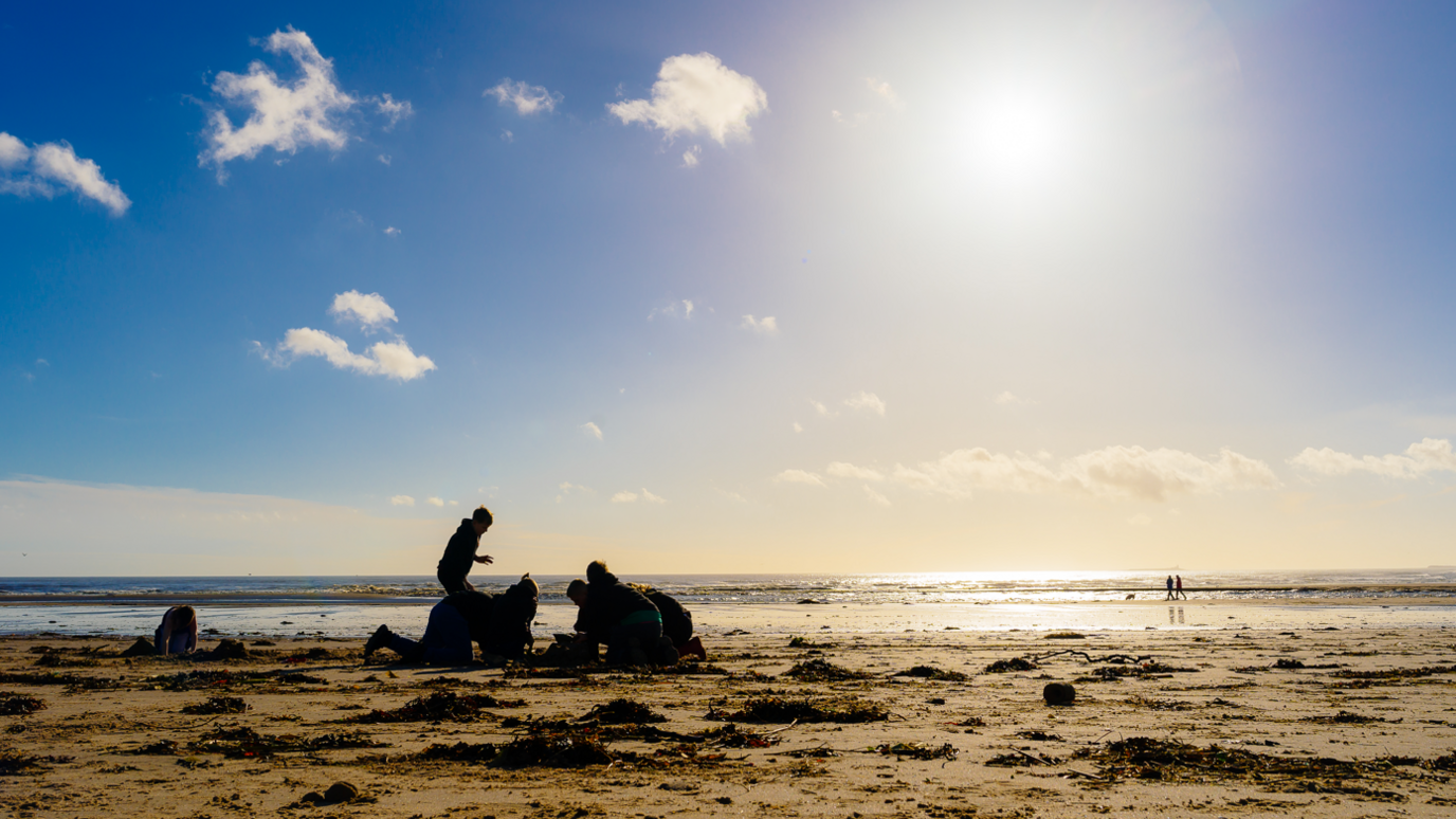 A group of children are building a sandcastle on a beach with the sea behind them. The sun is low in the sky so they are mostly black shapes with a stretch of sand behind lit by the suns rays. 