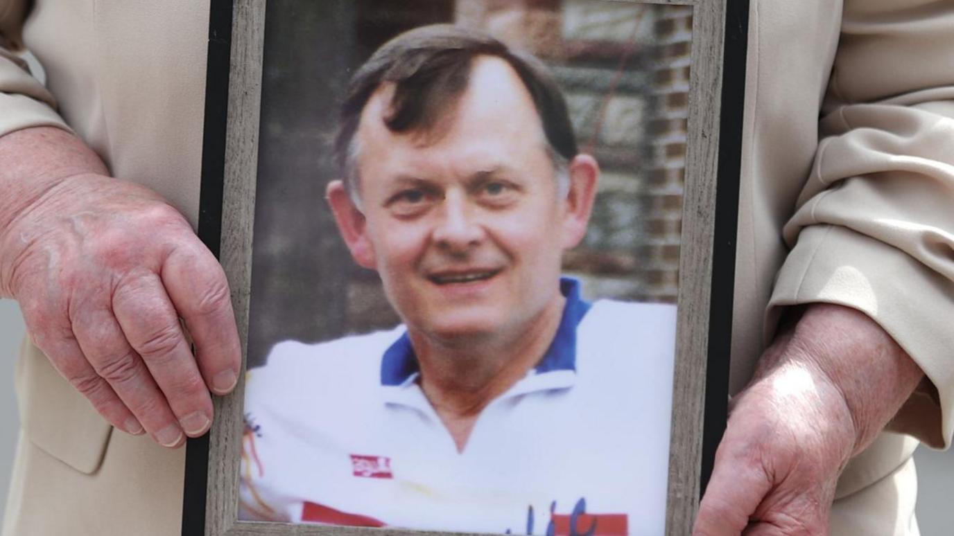 A close up image of a person holding a framed, colour photo of Sean Brown. Mr Brown has medium-length brown hair with some grey parts on the side of his head. He is smiling at the camera and is wearing a white polo shirt with blue and red designs on it.