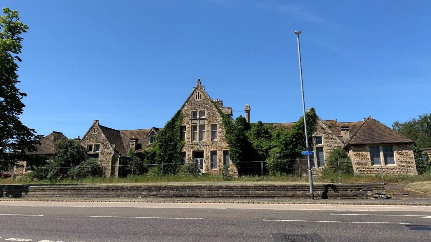 A disused old building with overgrown grass and greenery growing up the walls. A fence is lined up along the front. The sky is a deep blue in the background.