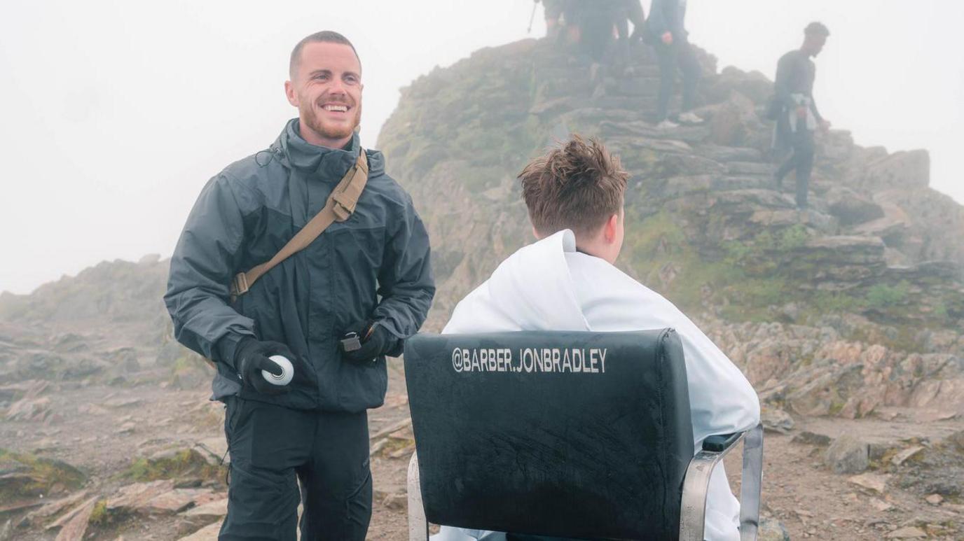 A smiling Jonathan Bradley wearing a black jacket and trousers at the top of a mountain. He is standing next to a barber chair, which a man is sitting in with a white towel around his shoulders and with his back to the camera.