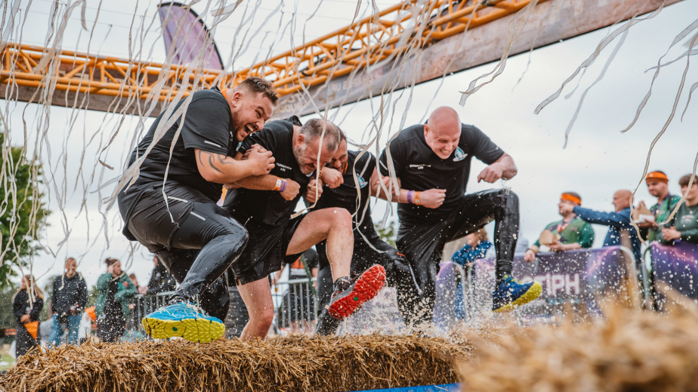 Four men in black t-shirts and leggings running through an obstacle together, with their heads down and arms linked, and all smiling. They are jumping over a hay bale with streamers falling down around them within a steel roof. There are spectators in the background