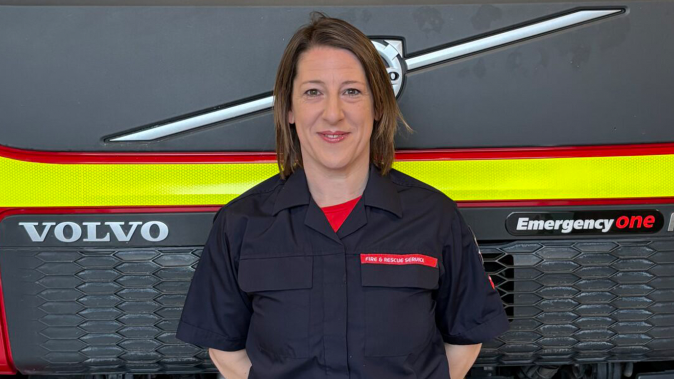 Carly Draper - A woman stands in front of a fire engine with her arms behind her back.