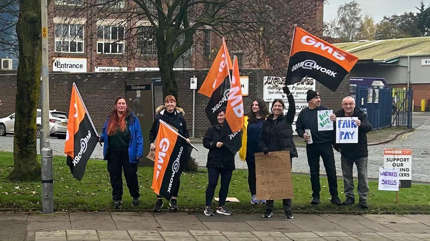 Seven people holding GMB orange flags and signs saying "Fair Pay". They are standing outside on a patch of grass and smiling. 