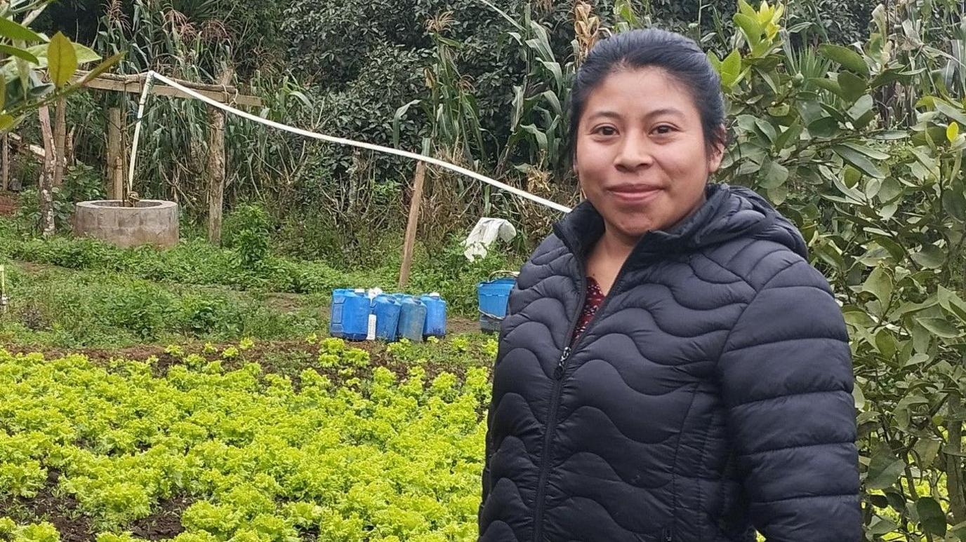 Farmer Sandra Noemi Bucu Saz smiles at the camera as she stands in front of her plot of land