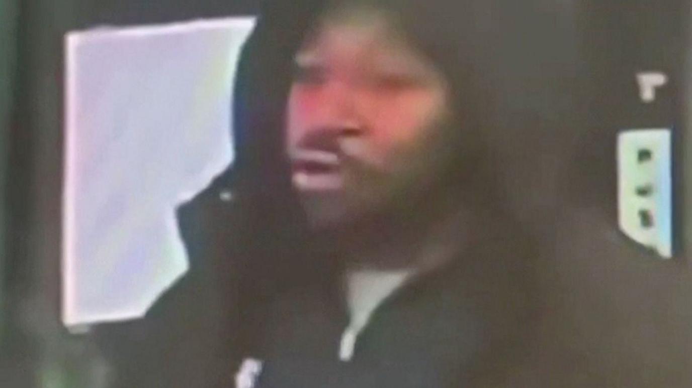 A man wearing a black hooded coat, pursing his lips, white standing inside.