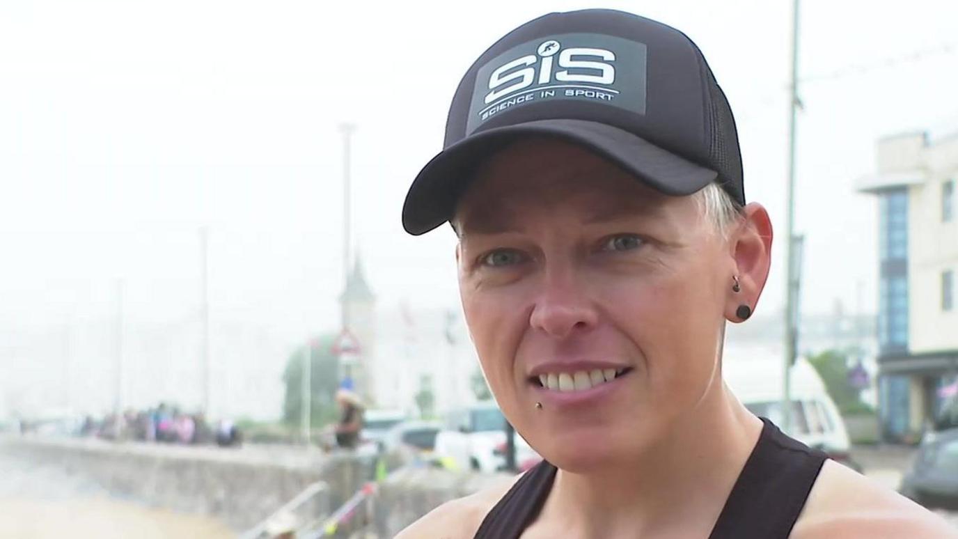 Lexi is wearing a black hat with the SiS logo and black vest top. She has blonde hair and piercings in her ears and below her mouth. She's smiling and is on Exmouth seafront. Buildings are behind her and you can see the beach in the foreground 
