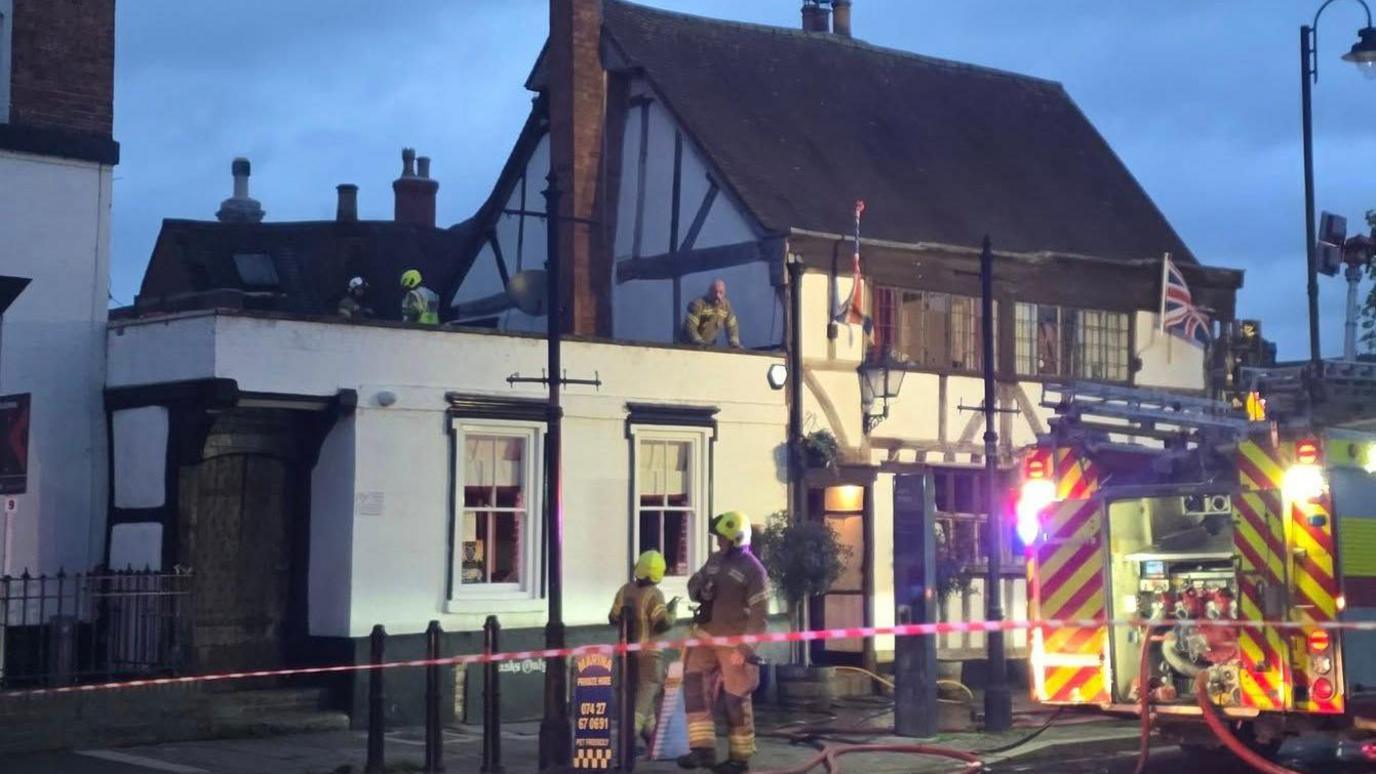 Firefighters at the Black Bear Pub. The road in front of the pub has been cordoned off with red tape and there is a fire engine parked on the road. There are two fire fighters outside of the pub and three on the roof.