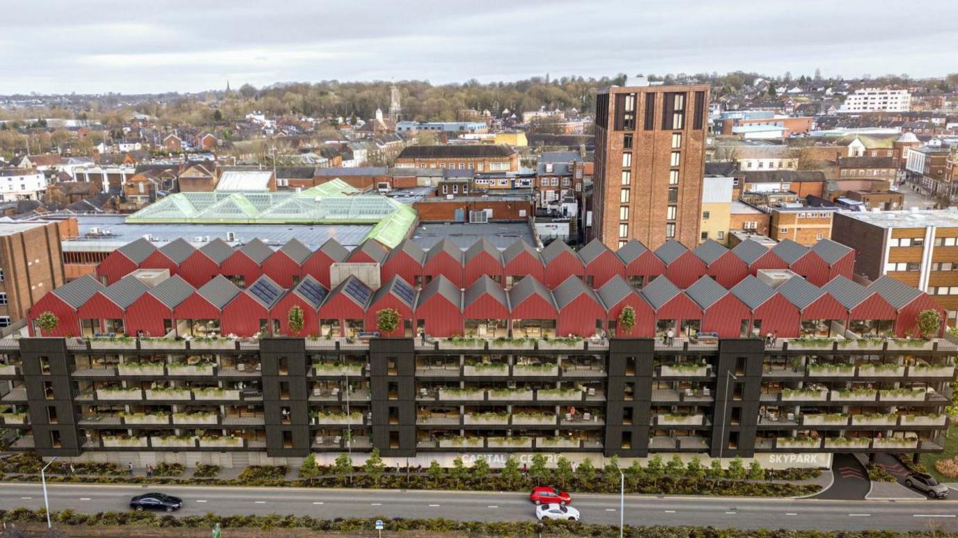 An aerial CGI showing the proposed flats - a large black building with balconies along its side and small red houses on the top. A main road is in front of the building with a townscape of Newcastle-under-Lyme in the background.