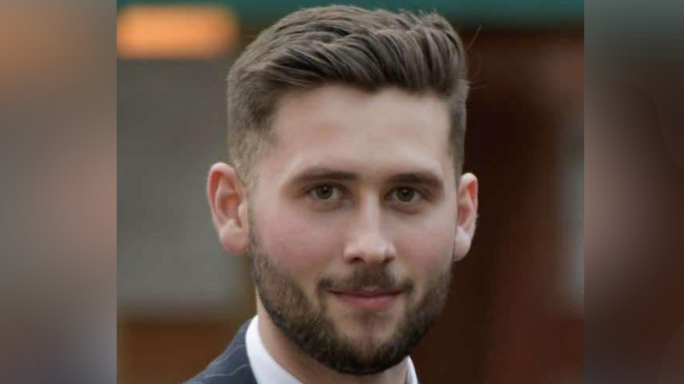 Rhys Askham smiles at the camera. He has short brown hair that has been styled. He has a dark coloured beard. He is wearing a suit with a white shirt. 