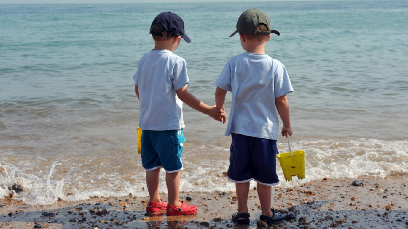 Two young boys hold hands as they stand at the water's edge on a beach. There are barely any waves and the water is clear. The boys are wearing white T-shirts and blue shorts with Crocs and caps. They are holding a bucket each.