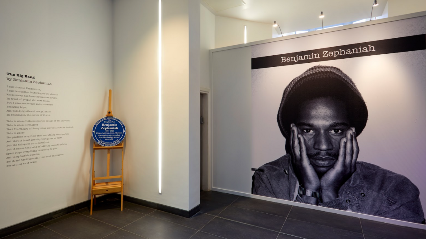 A black and white photo of Benjamin Zephaniah, which fills an entire wall, is next to a blue plaque resting on a wooden stand