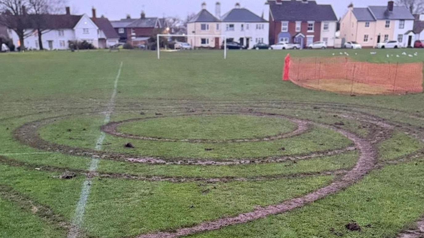 Circles of muddy tyre tracks left by a car, most likely doing doughnuts on the field. There is a football goal in the background and a row of houses behind it. Part of the pitch has already been fenced off by orange netting.