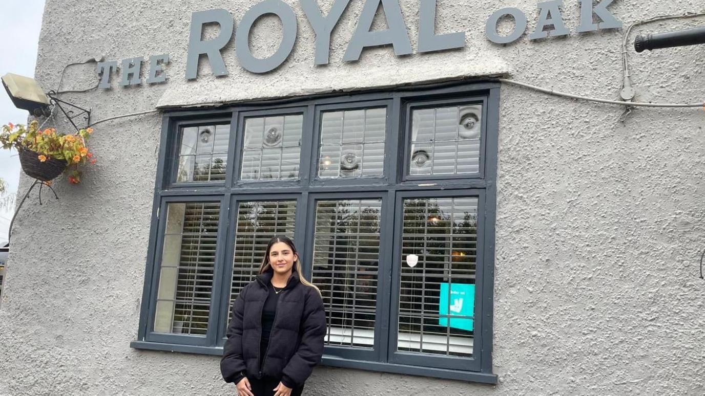 A woman with long black and blonde hair wearing black top, trousers and puffer jacket. She is stood outside a grey pub building with a lettered sign saying The Royal Oak.
