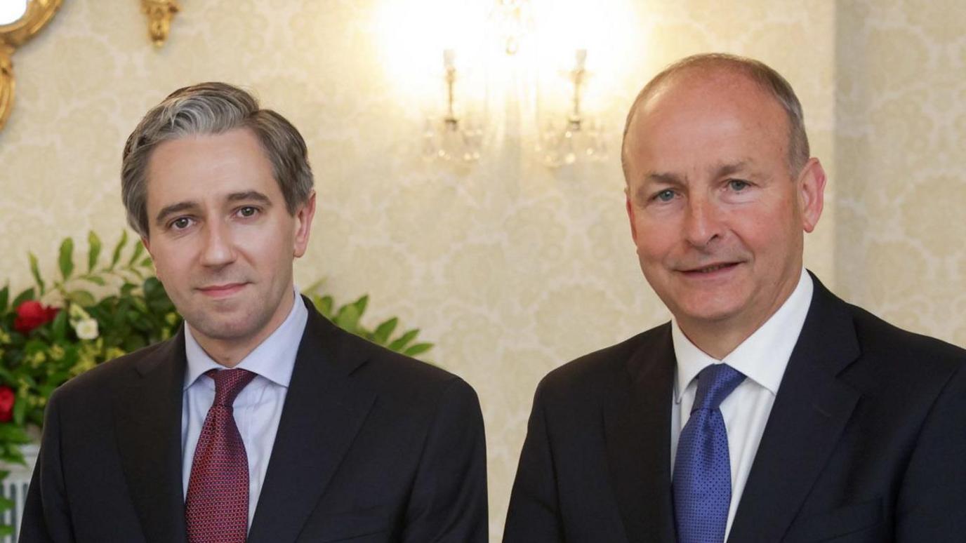 Simon Harris and Micheál Martin. Simon has grey-ish hair and wearing a black blazer, light blue shirt and burgundy tie. Micheál is balding and wearing a black blazer, white shirt and blue tie. Both are looking at the camera and a floral arrangement can be seen in the background. 