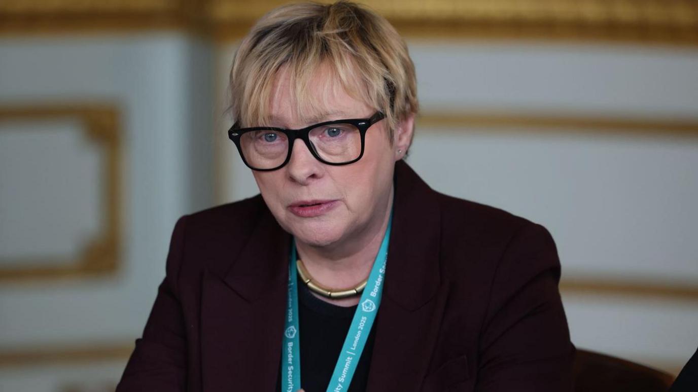 Labour MP Dame Angela Eagle has short blonde hair and is wearing a dark suit jacket and top and black-framed glasses. She is wearing a gold necklace and a light blue lanyard around her neck.
