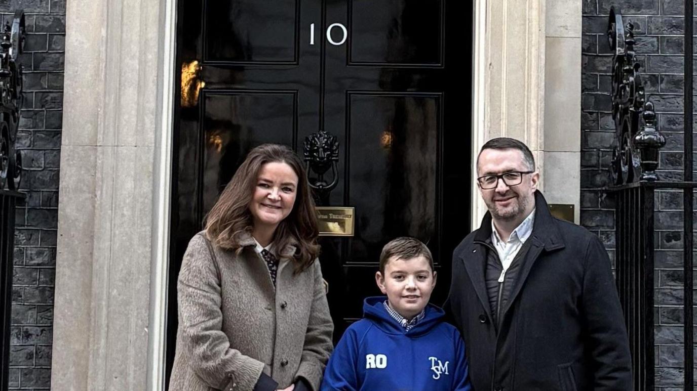 Roman stands in the middle of his dad Ryan and MP Jenny Riddell-Carpenter outside 10 Downing Street. Roman wears a blue hoodie, has short dark hair and holds a letter written on a piece of paper. Ryan has short dark hair, glasses and wears a black coat. Jenny has long dark hair and wears a long grey coat.