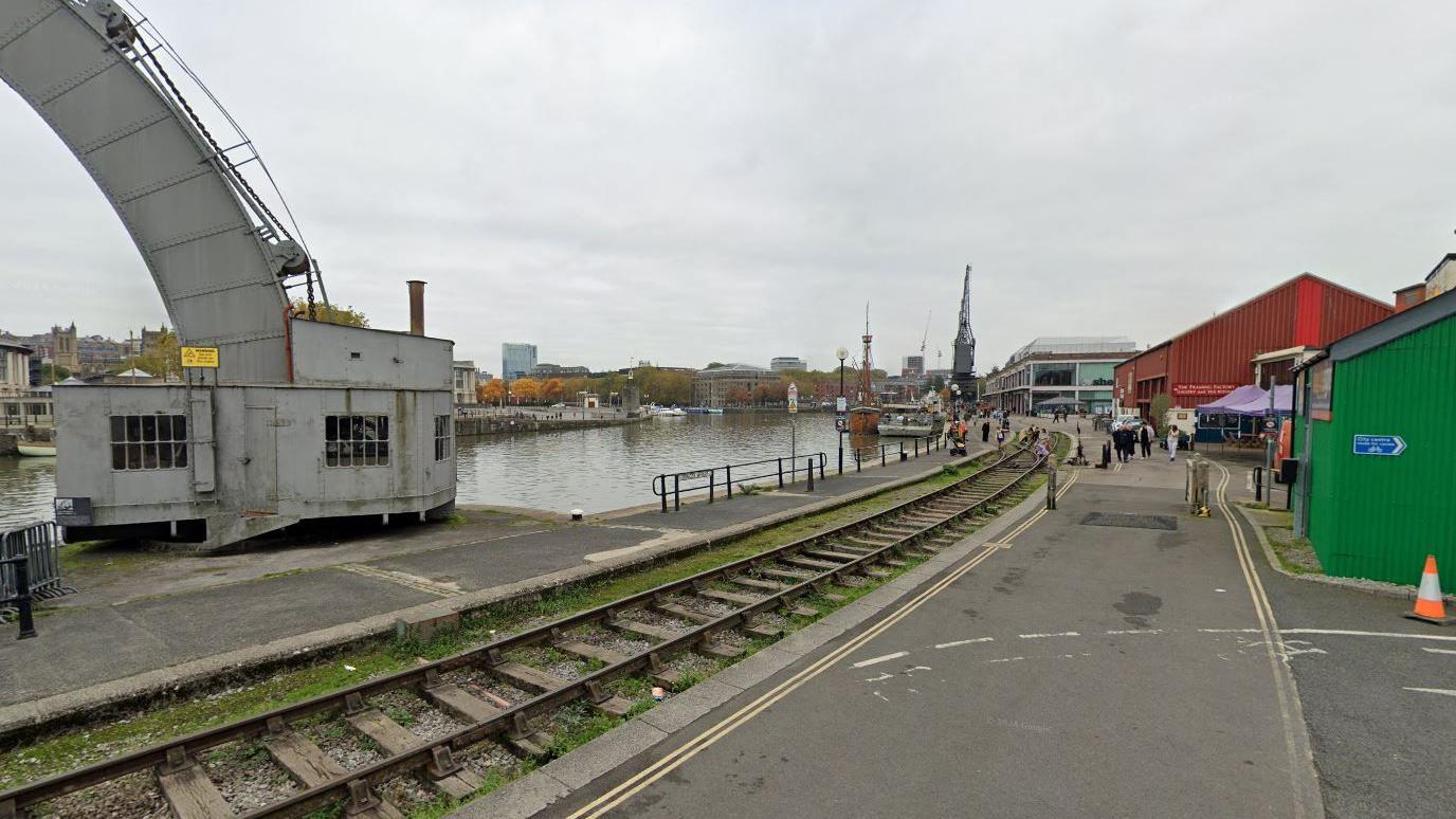 A google street view screenshot showing Wapping Wharf in Bristol's city centre. There is a disused railway line running through the pavement and the harbour is visible in the background. 