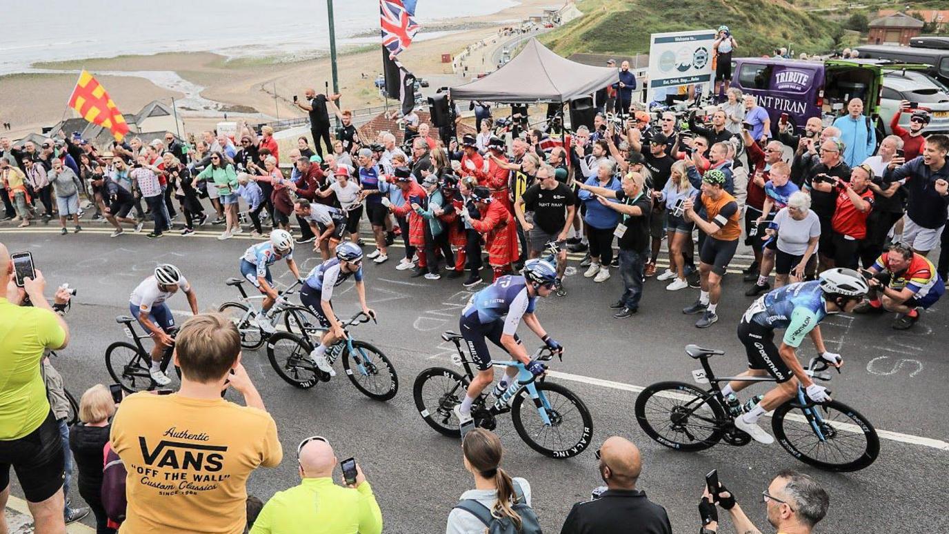 A line of five cyclists ascending a steep hill with a view of a beach below. On either side of the road are people waving and cheering