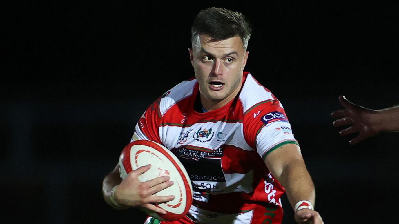 Ioan Hughes holds a rugby ball wearing a red and white hooped jersey