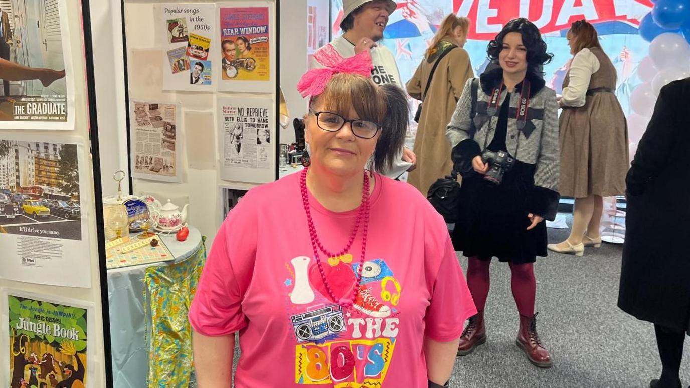 Joanne Clark is wearing a pink T-shirt which says "I love the 80s". She is inside a hallway. She has her hair in a ponytail and wears glasses and a pink necklace. Behind her is a woman with a black wig holding a Canon camera and a man in a bucket hat. There are other people further back. On the left hand side are photos and posters from old films and adverts. The window in the back says "VE Day".