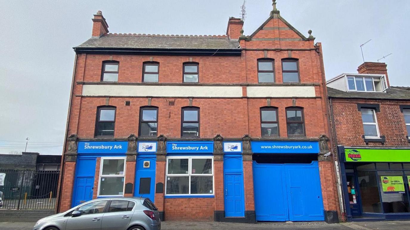 The front of the Shrewsbury Ark building. It is red brick with blue wooden doors and signs . There is a silver car parked outside. 